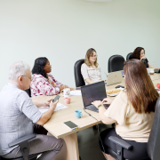 Foto de uma mesa de reuni&otilde;es com 6 pessoas sentadas, sendo 2 homens e 4 mulheres. Eles olham para dispositivos eletr&ocirc;nicos como celulares e laptops. Ao fundo, uma TV projeta a transmiss&atilde;o da sess&atilde;o, com pessoas participando virtualmente.