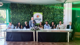 Foto colorida de uma mesa com 8 pessoas sentadas. Ao centro, um homem de terno est&aacute; em p&eacute; e fala ao microfone. Ao fundo, h&aacute; uma parede de heras artificiais e um banner com o logotipo da Uergs.