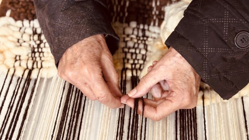 Foto colorida de m&atilde;os de uma mulher idosa vestindo casaco marrom, trabalhando com fios de l&atilde; de cores bege e marrom em um tear.