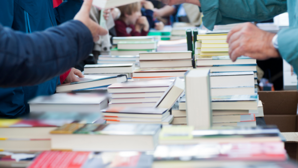 Bancada com in&uacute;meros livros para comerciliza&ccedil;&atilde;o com algumas pessoas ao redor, das quais aparecem apenas bra&ccedil;os e m&atilde;os.