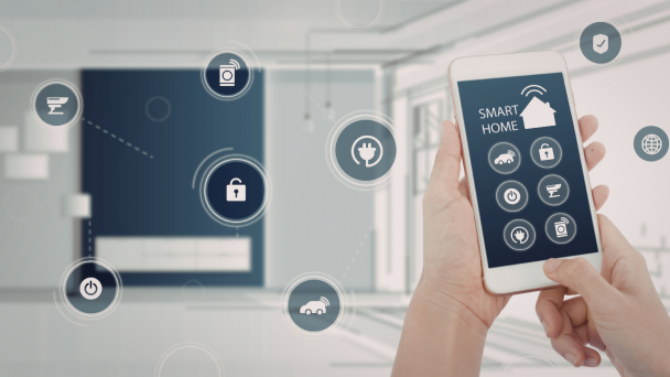 &Agrave; direita, m&atilde;os seguram um celular smatphone que tem na tela &iacute;cones que s&atilde;o de servi&ccedil;os relacionados &agrave; resid&ecirc;ncia e controlados pelo aparelho.