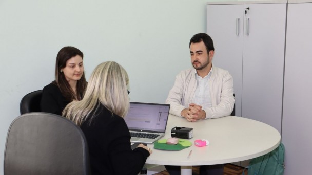 Tr&ecirc;s pessoas est&atilde;o reunidas em uma sala de escrit&oacute;rio, sentadas ao redor de uma mesa redonda branca. Em primeiro plano, uma mulher de cabelos loiros aparece de costas, utilizando um notebook aberto sobre a mesa. &Agrave; frente dela, um homem de cabelos escuros e barba curta, vestindo cal&ccedil;a cinza, camisa clara e jaqueta bege, est&aacute; sentado com as m&atilde;os apoiadas uma sobre a outra. Ao lado dele, uma mulher de cabelos castanhos, usando roupa escura, acompanha a conversa. Sobre a mesa h&aacute; materiais de escrit&oacute;rio, como canetas, marca-textos, mouse e um carimbo. Ao fundo, aparecem arm&aacute;rios brancos e uma mochila verde apoiada no ch&atilde;o. O ambiente tem paredes claras e piso de madeira em padr&atilde;o geom&eacute;trico.