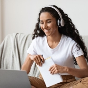 Fotografia horizontal de uma mulher sentada em um sof&aacute;, em um ambiente interno bem iluminado. Ela usa fones de ouvido, camiseta branca e cal&ccedil;a marrom, e sorri enquanto olha para um computador port&aacute;til apoiado &agrave; sua frente. Nas m&atilde;os, segura um caderno espiral e uma caneta. Ao fundo, h&aacute; uma planta em um vaso, &agrave; esquerda, e parte de um m&oacute;vel ou objeto decorativo, &agrave; direita.