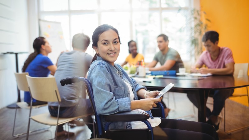 Foto de uma mulher morena clara de jaqueta jeans, segurando um tablet, sentada em uma cadeira de rodas e sorrindo. Ao fundo, uma mesa com um grupo de estudantes no entorno.